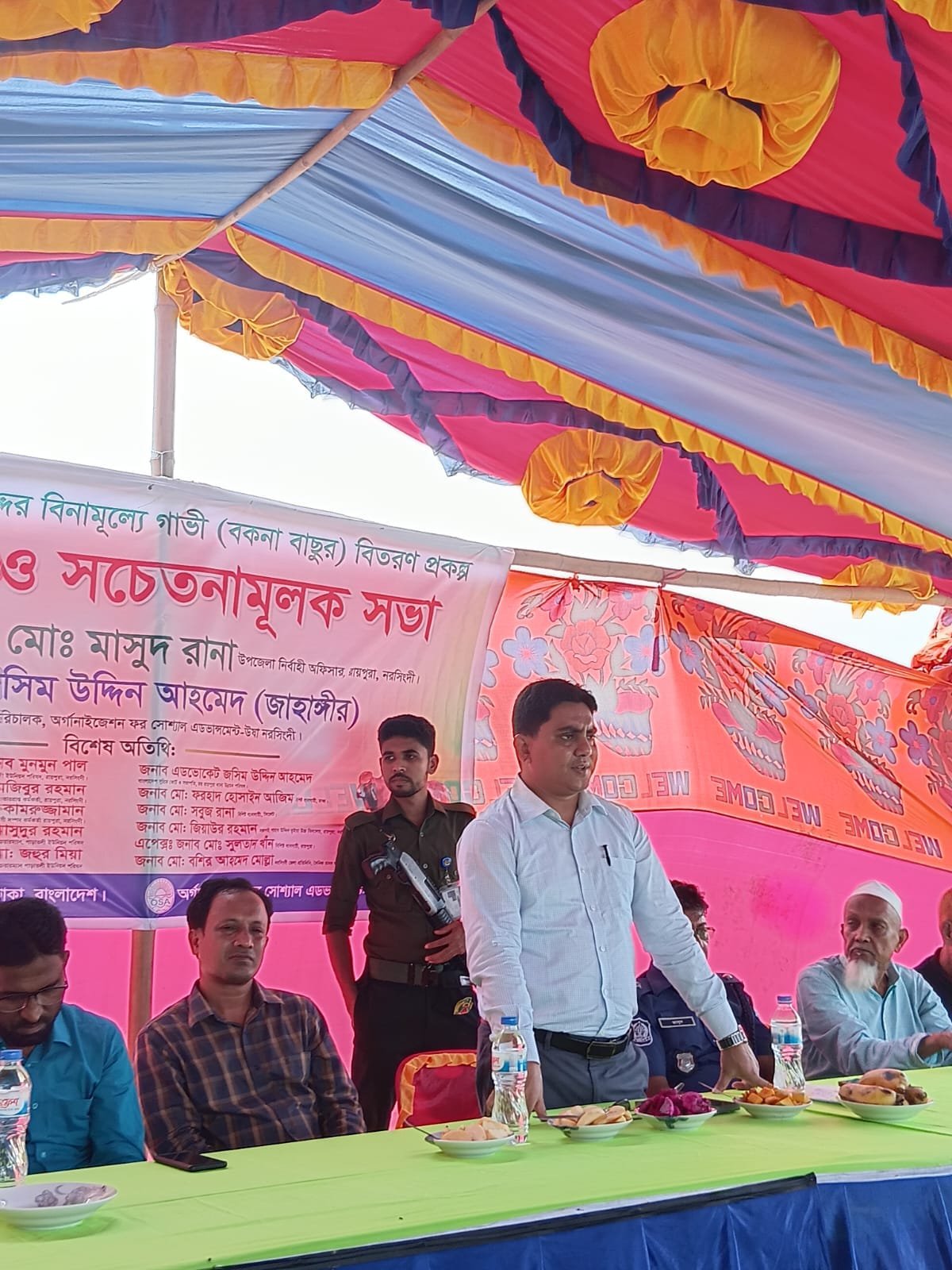 A guest addressing the awareness meeting panel, tent drapes and a floral screen behind him.
